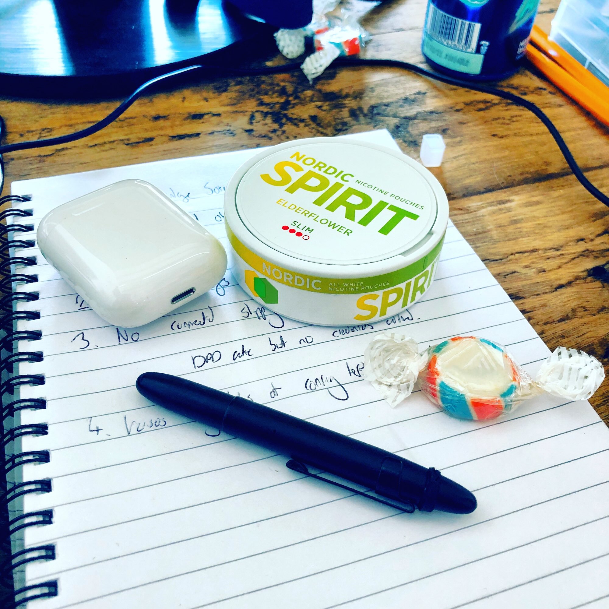 A can of Nordic Spirit nicctine pouches on a notepad with pad, AirPods and a sweet.